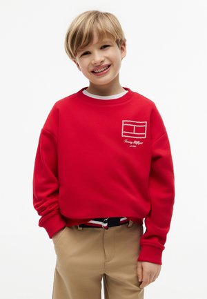Uśmiechnięte dziecko o blond włosach, ubrane w czerwoną bluzę Tommy Hilfiger i beżowe spodnie, stojące z rękami w kieszeniach.