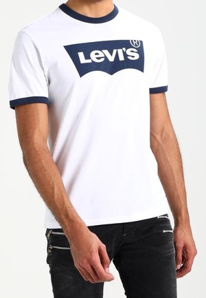 Homme portant un t-shirt blanc Levi's avec un col et des bordures de manches bleu marine, associé à un pantalon noir avec des détails de fermeture éclair.