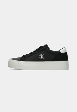 Black sneakers with a sleek design, featuring a white heel accent, textured fabric, round toe, and prominent logo on the side. Platform sole.