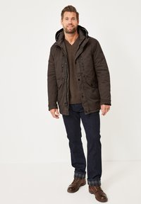 Braune Jacke mit Kapuze, ausgestattet mit Reißverschlusstaschen und einem gesteppten Muster, kombiniert mit dunklen Jeans und braunen Schnürstiefeln.