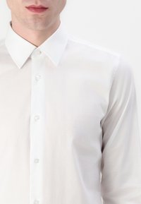 Biela košeľa s dlhým rukávom a špicatým golierom, s jemným leskom. Má gombíkovú prednú časť a priliehavý strih.