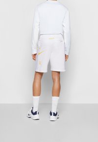 Bílé sportovní šortky s bočními panely z síťoviny a žlutým logem Nike, spárované se světle modrou dlouhýrukávovou košilí a bílými ponožkami, nošeny s botami.