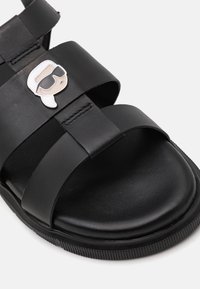 Musta nahkasandaali, jossa on leveät remmit, teksturoitu pohjallinen ja huomattava leikkisä hahmopinssi ylärimassa. Kumipohja, jossa on uria.