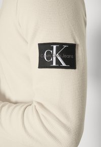 Beige långärmad skjorta med ett texturerat mönster. Har en svart lapp på ärmen med vit text: "Calvin Klein Jeans" och en "CK"-logotyp.