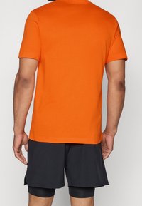 Camiseta de manga corta con cuello redondo en algodón naranja brillante, mostrada desde la parte de atrás, combinada con pantalones cortos negros. Diseño liso, sin patrones visibles.