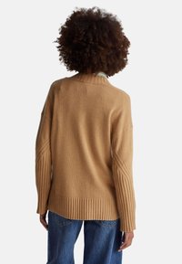 Hellbrauner Strickpullover mit hohem Kragen, gerippten Bündchen und Saum sowie detaillierter Nahtführung an den Ärmeln. Trägt man über blauen Jeans.