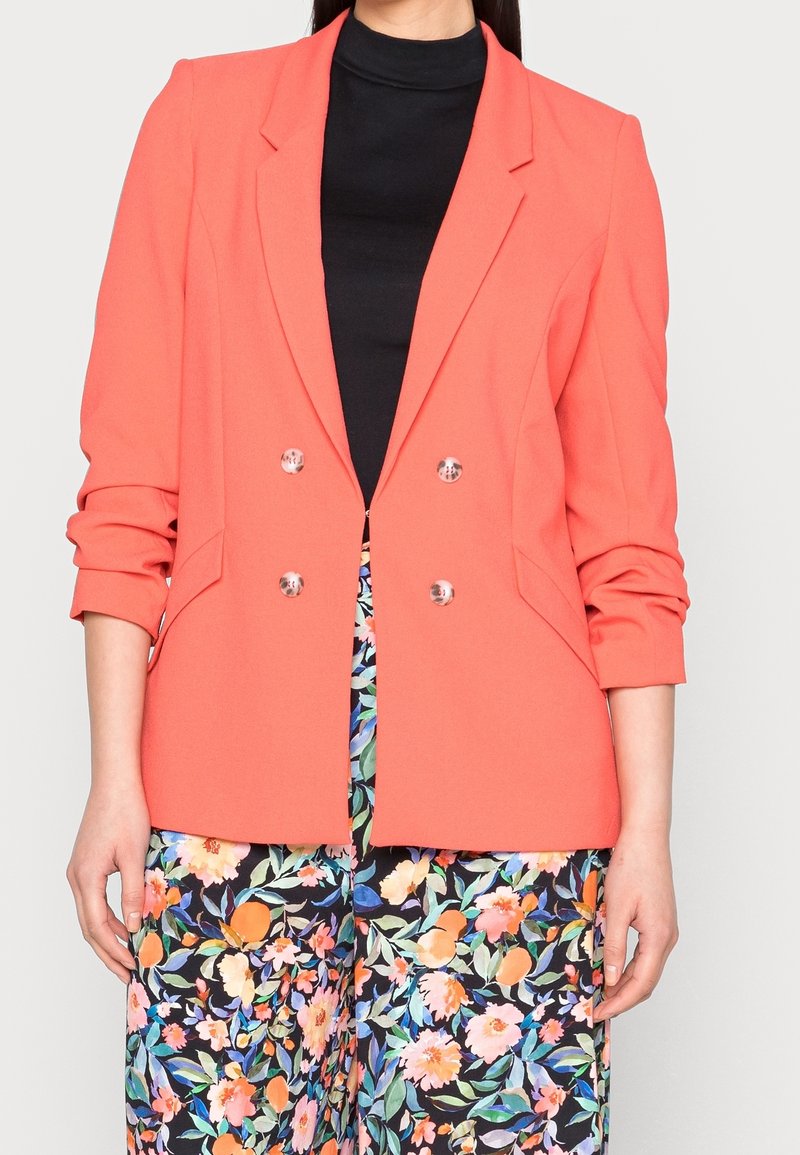 Femme portant un blazer corail à double boutonnage, un haut noir et un pantalon à motif floral aux couleurs rose, orange et vert.