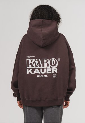 Person mit lockigen Haaren, von hinten gesehen, trägt einen dunkelbraunen Hoodie mit großem weißen "KARO KAUER" Text auf dem Rücken.