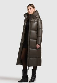 Lange pufferjas in donkerolijf groen, met een hoge kraag, gestikte ontwerpen en ritssluiting. Model draagt enkelhoge zwarte laarzen.