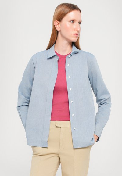Camisa de botones azul claro con cuello, hecha de una tela suave. Llevada sobre una camiseta rosa y pantalones beige, con un corte relajado y mangas largas.