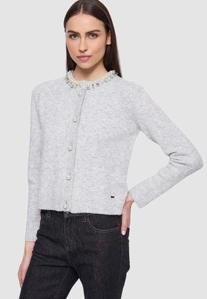 Donna che indossa un cardigan grigio chiaro con perle e gioielli a decorare il colletto e bottoni perlacei, abbinato a jeans neri.