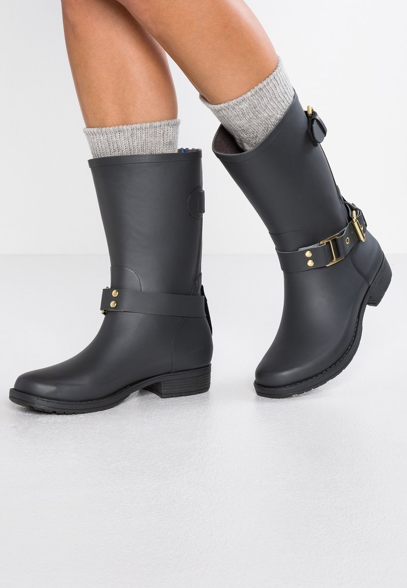 Botas de goma negras con una superficie texturada, detalle de hebilla y correa en el tobillo. Calcetines de lana gris son visibles por encima de las botas. Suela plana.