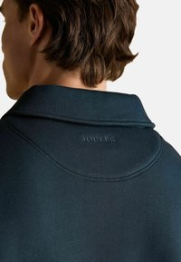 Vīrietis valkā tumši zilu krekla augšējo daļu ar vārdu "Joules" izšūtu augšējā aizmugurē, redzams no aizmugures uz vienkrāsaina fona.