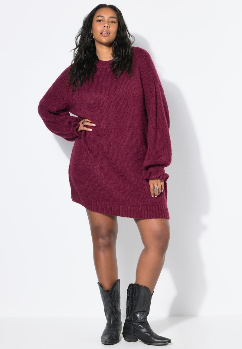 Burgunderfarbene, gestrickte Oversize-Sweatkleid mit geripptem Saum, langen Puffärmeln und lockerer Passform. Tragen mit schwarzen Cowboy-Stiefeln.