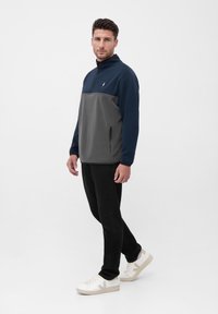Fleece-Jacke mit Viertelreißverschluss, navyblauem Oberteil und grauem unteren Abschnitt, seitlichen Reißverschlüssen und Logodetail. Kombiniert mit schwarzen Hosen und weißen Turnschuhen.