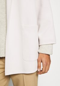 Main portant un pantalon beige et un pull en maille crème, couverte d'une veste blanche texturée avec une poche visible et une manche retroussée.