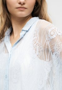 Close-up of a woman wearing a light blue lace blouse with floral patterns and a collared neckline.