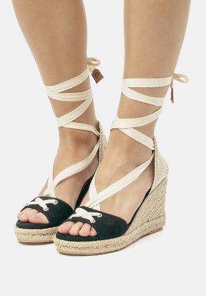 Wedge espadrilles nere e beige con cinturini incrociati in tessuto, punta aperta e suola intrecciata in corda. Presenta dettagli in pizzo bianco sulla punta.