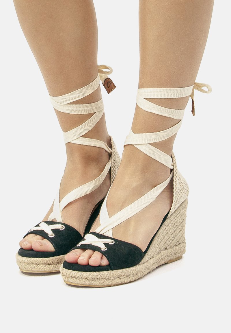 Schwarze und beige Espadrille-Wedges mit gekreuzten Stoffriemen, offener Zehenpartie und aus Seil gewebter Sohle. Mit weißen Spitzenverzierungen an der Zehenpartie.