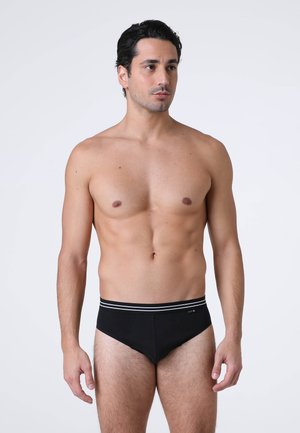 Uomo con capelli corti e scuri che indossa mutande nere con una fascia a righe in vita, in piedi davanti a uno sfondo bianco uniforme, che guarda di lato.