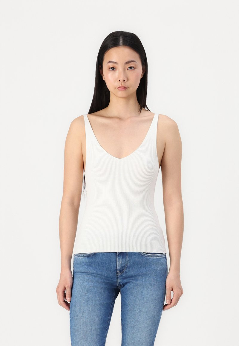 Top a maniche sottili bianco a coste con scollatura a V profonda, abbinato a jeans in denim blu. Il tessuto ha un aspetto liscio e texture.