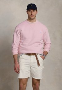 Világos rózsaszín pulóver, fehér farmer rövidnadrág tépett széllel, barna fonott öv, és sötétkék baseball sapka logóval. Minimális minták, laza design.