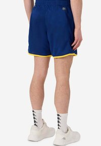 Pantaloni sportivi della marina con rifiniture gialle, vita elastica e dettaglio del logo. Indossati con sneakers bianche e calzini a fantasia.