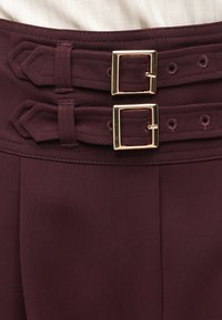 Κοντινό πλάνο σε μπορντό παντελόνι με διπλές ζώνες από ύφασμα, που διαθέτουν χρυσές, τετράγωνες αγκράφες και ραμμένους θηλιούς ζώνης στη μέση.