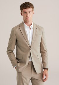 Jeune homme portant un costume beige avec une chemise blanche, les mains détendues, se tenant contre un fond uni beige clair.