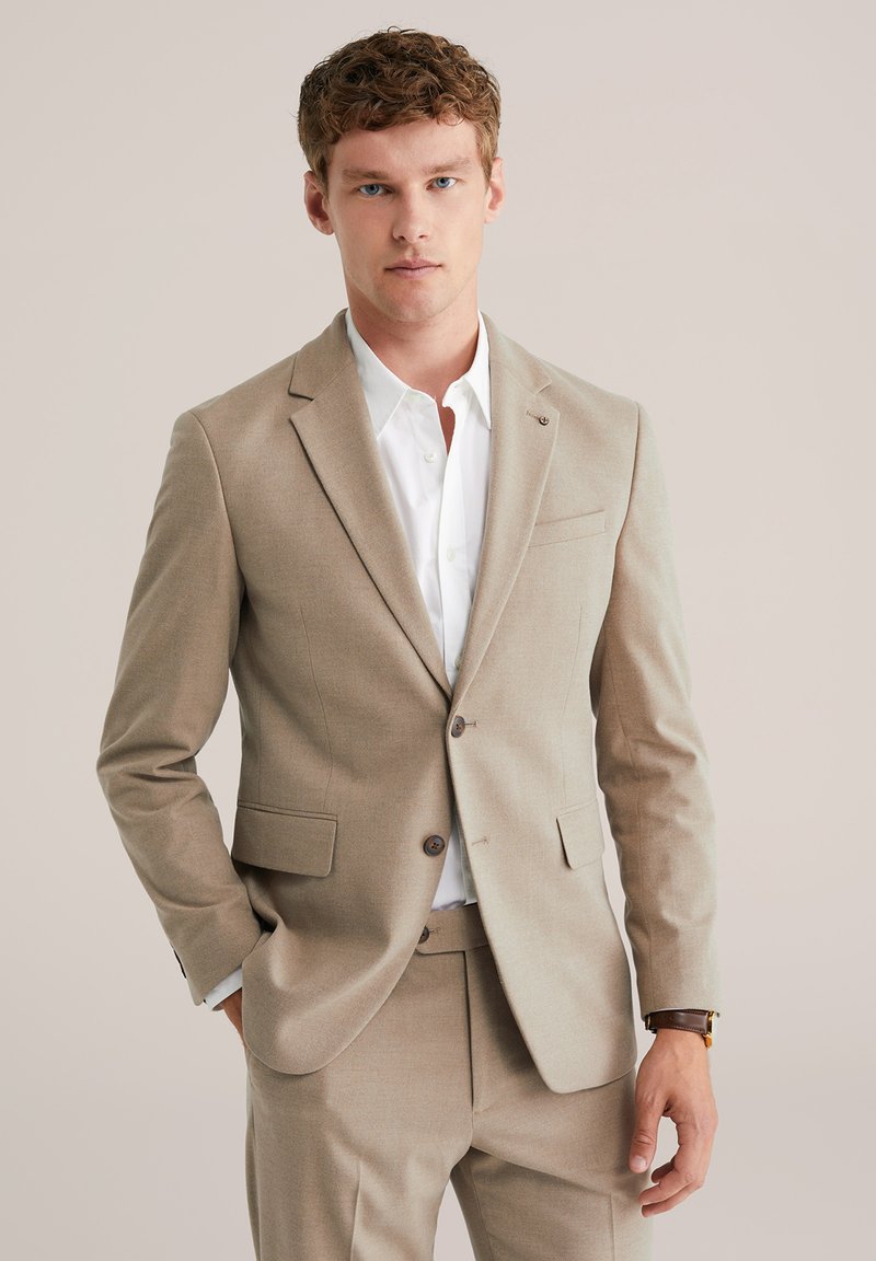 Jeune homme portant un costume beige avec une chemise blanche, les mains détendues, se tenant contre un fond uni beige clair.