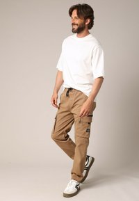 Pantalon cargo beige avec des poignets élastiques, plusieurs poches et une taille élastiquée avec cordon, associé à un T-shirt blanc uni et des baskets blanches.