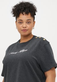 Szara bawełniana koszulka z okrągłym dekoltem i krótkimi rękawami, ozdobiona złotymi guzikami na jednym ramieniu oraz haftowanym tekstem.