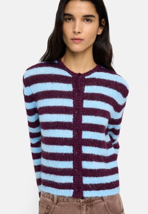 Kobieta ubrana w zapinany na guziki, puszysty sweter z poziomymi paskami w jasnoniebieskim i ciemnofioletowym kolorze, zestawiony z brązowymi spodniami.