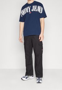 Man som bär en marinblå Tommy Jeans t-shirt, svarta cargobyxor med logotypplåster och vit-blå sneakers, står mot en enfärgad bakgrund.