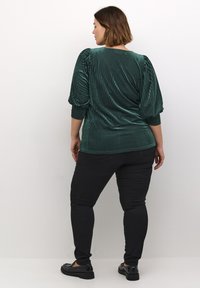 Grøn fløjlsbluse med vertikale striber, puffede ærmer og ribbede manchetter, parret med sorte slim-fit bukser og sorte sko.