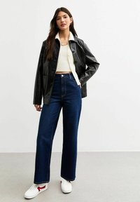 Zwarte leren jas over een witte top, gecombineerd met donkerblauwe wijde jeans en witte sneakers met rode accenten. Gladde textuur, moderne pasvorm.