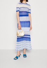 Robe en maille avec un motif en zigzag dans des nuances de bleu et blanc, manches courtes et corsage ajusté. Associée à un sac en satin doré et des sandales turquoise.