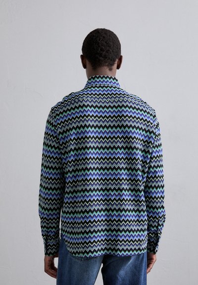 Μακρυμάνικη μπλούζα με μαύρη βάση, διακοσμημένη με σχέδιο ζιγκ ζαγκ σε μπλε και πράσινο. Το ύφασμα φαίνεται λείο με κλασικό γιακά και λεπτομέρειες από κουμπιά.