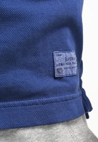 Blå texturerad tyg med en vävd yta, prydd med en sydd etikett med vit text. Design av bomullsblandning, med grå kant som syns nedanför.