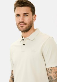 Beige poloshirt med struktureret stof, to sorte knapper ved kraven og et diskret logo på venstre bryst. Korte ærmer.