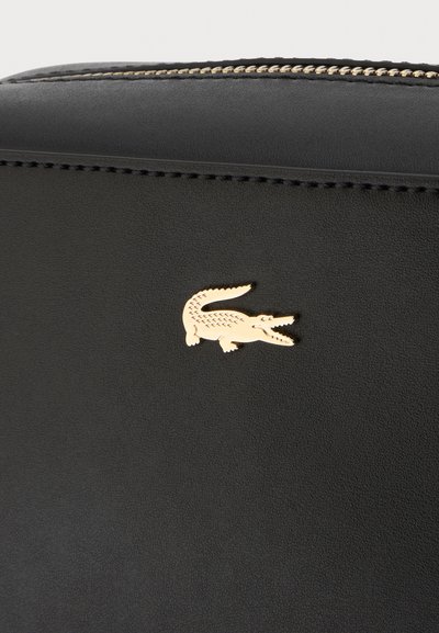 Surface en cuir noir avec des coutures visibles et un petit logo doré en forme de crocodile près du centre supérieur sous une fermeture éclair.