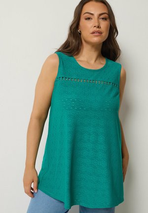 Frau trägt ein ärmelloses türkisfarbenes Oberteil mit Lochstickerei im Blumenmuster und horizontalem Spitzenbesatz über der Brust, kombiniert mit hellblauen Hosen.