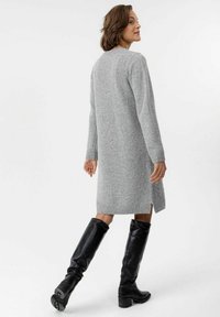 Graues Strickkleid mit langen Ärmeln, gerippten Bündchen, seitlichen Schlitzen und einem lockeren Schnitt. Getragen mit hohen schwarzen Lederstiefeln.