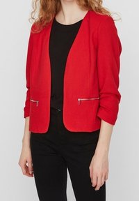 Red tailored blazer with long sleeves, textured fabric, open front, and silver zippered pockets. Worn over a black top and dark pants.