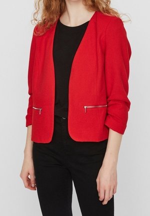 Rode op maat gemaakte blazer met lange mouwen, textuurstof, open voorkant en zilveren ritszakken. Draagt over een zwarte top en donkere broek.