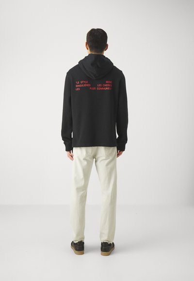 Sweatshirt à capuche noir avec texte rouge au dos, associé à un pantalon crème et des baskets sombres. Tissu doux, coupe décontractée.