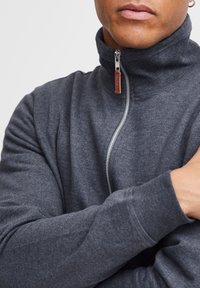 Sudadera con cremallera en gris oscuro, con cuello alto, puños acanalados y una cremallera metálica que presenta una etiqueta de cuero marrón. Textura suave, diseño ajustado.