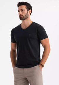 Czarny t-shirt z dekoltem w serek wykonany z miękkiego materiału, z krótkimi rękawami i małym, wytłoczonym logo na piersiach, w połączeniu z jasnobeżowymi spodniami.