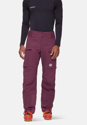 Mann trägt lila Outdoor-Hosen mit Reißverschlusstaschen am Oberschenkel und orangefarbene Bergstiefel, kombiniert mit einem schwarzen Langarmshirt.