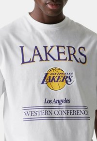 T-shirt bianco in cotone con il testo audace in viola "LAKERS" e "LOS ANGELES", accompagnato da una grafica di un pallone da basket giallo, e con "WESTERN CONFERENCE" sotto.